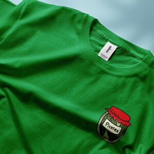 May include: A green t-shirt with a graphic of a jar of pickles. The jar has a red lid and the word "Pickles" is printed on the label.