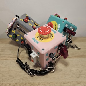 Pode incluir: Três cubos sensoriais coloridos com vários botões, interruptores e luzes. O cubo rosa tem um grande botão de parada vermelho e a palavra "STOP" em amarelo. O cubo cinza tem um controle deslizante e o cubo azul turquesa a palavra "EMERGENCY".