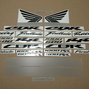 May include: A collection of black and white adhesive decals for a motorcycle, featuring the text CBR, Fireblade, Racing, 1000, 600, and RR, along with two wing-shaped graphics and several plain rectangular strips for decorative application.