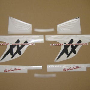 May include: A set of motorcycle decals featuring two large white wing graphics, two CBR logos, two 1100 Super Blackbird emblems with black XX lettering, and two Dual CBS Evolution decals in red and white text, all arranged on a plain brown surface.