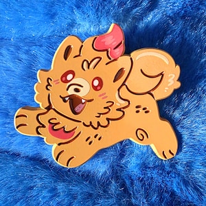 May include: A light orange enamel pin in the shape of a cartoon dog, running with a happy expression. The pin has red eyes, a pink tongue and ear, and is set against a blue, fluffy background.