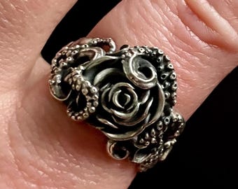 May include: A silver ring featuring a rose design at the center, surrounded by tentacles. The ring is worn on a finger, with another ring visible in the background. The ring has a detailed, gothic style.