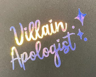 May include: A black rectangular card featuring the text Villain Apologist in a holographic, iridescent foil finish. The lettering is in a cursive script, accompanied by two four-pointed star icons and two small dots to the right of the text.