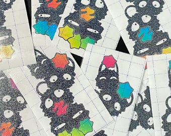 May include: A collection of rectangular stickers featuring a vertical stack of three black, glittery, cartoon-like creatures with large white eyes. Each sticker includes colorful, holographic star-shaped accents in shades of pink, blue, and green.