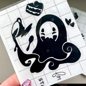 May include: Black vinyl stickers featuring a cartoon ghost-like character holding a spoon and teapot, with a slice of cake and hearts. The stickers are on a grid background.