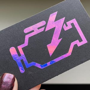Może przedstawiać: Czarna wizytówka z holograficznym symbolem kontrolki silnika. Symbol jest obrysowany na różowo i fioletowo, z błyskawicą pośrodku. Karta jest trzymana przez osobę.