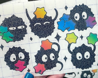 May include: A rectangular sticker sheet featuring six dark blue glittery soot sprite characters with large white eyes. The sprites are accented with colorful, gradient-colored star and leaf shapes in shades of pink, blue, green, and yellow.