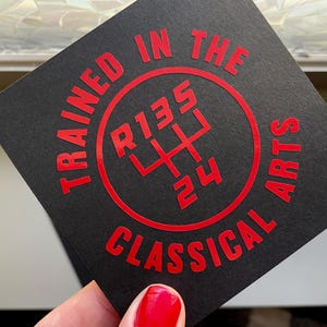May include: Black card with red text that reads "Trained in the Classical Arts" and a red circle with a stylized gear shift pattern inside. The gear shift pattern shows the letters "R135" and the numbers "24".