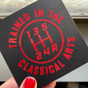 May include: Black sticker with red text and a red gear shift pattern. The text reads "Trained in the Classical Arts".