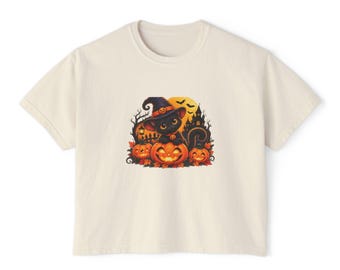 May include: A cream-coloured short-sleeve t-shirt featuring a graphic print of a black cat wearing a witch hat, sitting on a large orange jack-o'-lantern. The scene includes smaller pumpkins, autumn leaves, a dark castle silhouette, and bats.