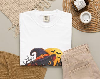 May include: A white t-shirt featuring a graphic of a black cat wearing a purple witch hat with an orange band and a small pumpkin accent, set against a large yellow moon, silhouettes of bare trees, flying bats, and a dark castle.