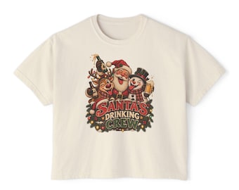 May include: A cream-coloured short-sleeve t-shirt featuring a graphic of Santa Claus, a reindeer, and a snowman holding drinks. The text "SANTA'S DRINKING CREW" is printed below the characters, surrounded by festive pine branches and lights.