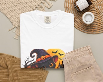 May include: A white crew neck t-shirt featuring a graphic of a black cat wearing a purple witch hat with an orange pumpkin accent, set against a yellow full moon, silhouetted trees, bats, and a house, with a Comfort Colors brand label.