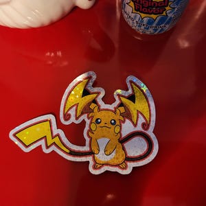 Puede incluir: Una pegatina holográfica con un personaje de dibujos animados amarillo con detalles de rayos. El personaje tiene una cola larga y está sobre un fondo rojo. La pegatina es probablemente un artículo de colección.