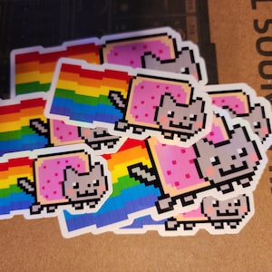Puede incluir: Una colección de pegatinas de Nyan Cat. Cada pegatina presenta un gato gris pixelado con un cuerpo rosa Pop-Tart, volando a través de un rastro de arcoíris. Las pegatinas están sobre una superficie marrón.