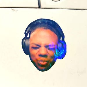 May include: A round sticker featuring a person wearing black headphones. The person's face is visible, with closed eyes and a slight expression. The sticker is affixed to a white car bumper, with a green grassy background.