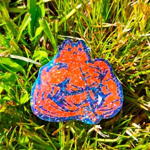 May include: A colorful, glittery resin pin featuring a muscular figure in shades of orange and blue. The pin is set against a backdrop of green grass, creating a vibrant contrast. The design is detailed and eye-catching.