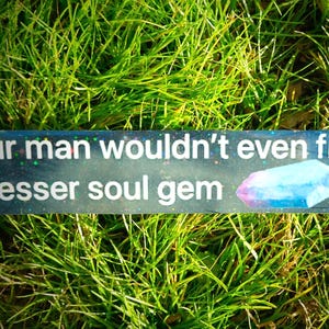 May include: A rectangular sign with the text "ur man wouldn't even fill a lesser soul gem" and a crystal illustration. The sign is set against a backdrop of green grass.