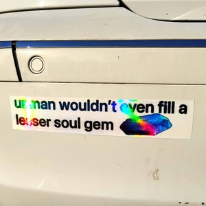 May include: A white bumper sticker with the text "ur man wouldn't even fill a lesser soul gem" in a holographic font. A colorful, gem-like graphic is below the text.