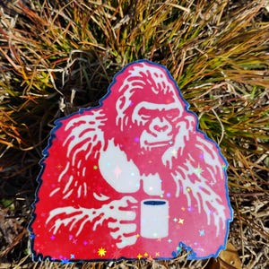 May include: A red sticker featuring a gorilla holding a mug. The gorilla is white with red accents and a blue outline. The sticker has a holographic, glittery effect with star-shaped sparkles.