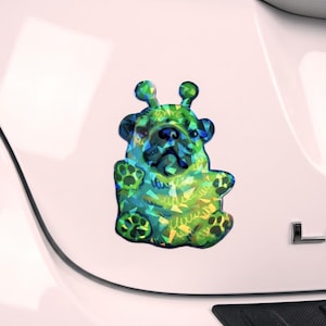 May include: A colorful sticker featuring a whimsical dog design with a unique alien-like appearance. The sticker showcases a vibrant mix of green, blue, and yellow hues, creating a playful and eye-catching aesthetic. The dog's features are stylized, with large eyes and a friendly expression.