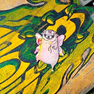 Pode incluir: Um autocolante extravagante com um pug de desenho animado vestido de fada. O pug tem asas cor-de-rosa, uma coroa de flores e segura uma varinha em forma de estrela. O autocolante est&aacute; sobre um fundo de arte abstrata amarela e verde.
