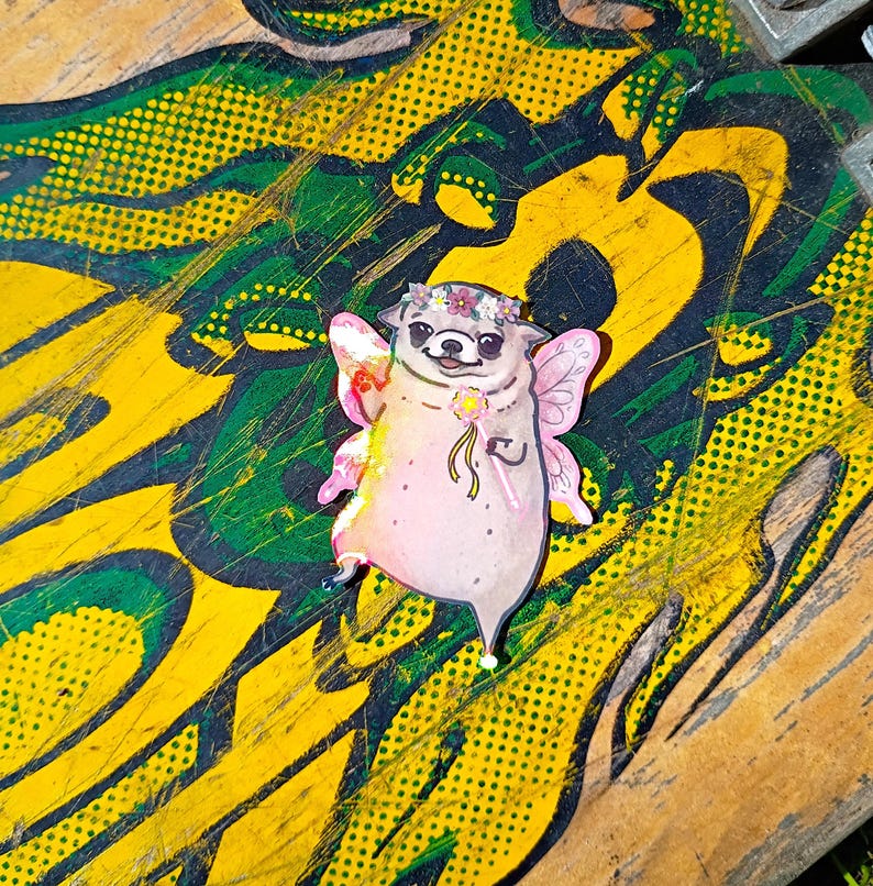 Pode incluir: Um autocolante extravagante com um pug de desenho animado vestido de fada. O pug tem asas cor-de-rosa, uma coroa de flores e segura uma varinha em forma de estrela. O autocolante est&aacute; sobre um fundo de arte abstrata amarela e verde.