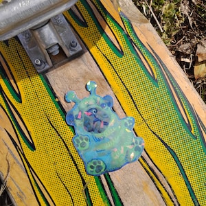 May include: A skateboard with a yellow and green design, featuring a blue sticker of a cartoon creature. The skateboard has a silver truck and wheels. The sticker is blue with pink accents and paw prints.