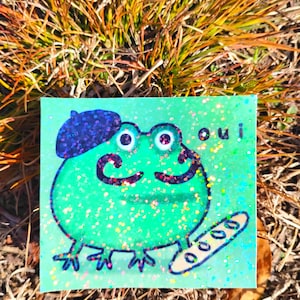 May include: A whimsical illustration of a green frog wearing a beret and a mustache, holding a baguette. The frog has large eyes and the word "oui" is written above it. The background is a glittery green.