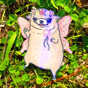 Pode incluir: Ilustra&ccedil;&atilde;o fantasiosa de um pug vestido de fada, completo com asas cor-de-rosa, uma coroa de flores e uma varinha m&aacute;gica. A obra de arte &eacute; ambientada em um fundo de grama verde e trevos.