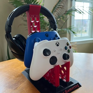May include: A 3D printed stand for video game controllers and headphones. The stand is black with red accents and has a slot for two controllers and a hook for headphones. The stand has the text "Your Gamertag" printed on the base.