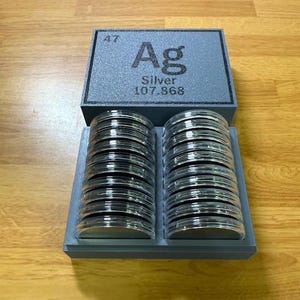 May include: A gray storage box with the periodic symbol for silver, Ag, and the atomic weight 107.868. The box holds two stacks of silver rounds.