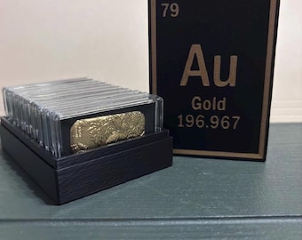 May include: A stack of gold bars in clear protective cases housed in a black box, accompanied by a black rectangular display card featuring the periodic table element 79, Au, Gold, and the atomic mass 196.967 in gold-colored lettering.
