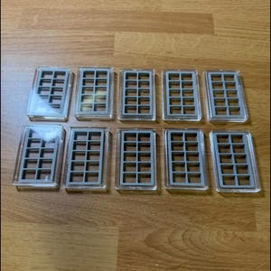 May include: Ten clear plastic coin holders with gray interior grids are arranged on a wooden surface. Each holder has a rectangular shape with a grid pattern inside, designed to hold and display coins. The holders are neatly organized.