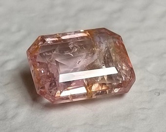 May include: A faceted, rectangular gemstone with a soft pink hue. The gem has a slight orange tint on some edges and is cut with multiple facets to reflect light. The gemstone is likely used in jewelry.