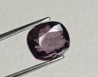 May include: A cushion-cut, purplish-gray gemstone is held by a pair of silver tweezers. The gemstone has a polished surface with facets that reflect light. The background is a plain, off-white color. This image is suitable for jewelry listings.