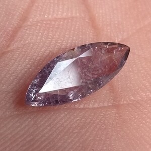 Puede incluir: Una gema de talla marquesa con un tono púrpura pálido. La gema es alargada y puntiaguda en ambos extremos, con inclusiones internas visibles. La piedra descansa sobre una superficie color piel.