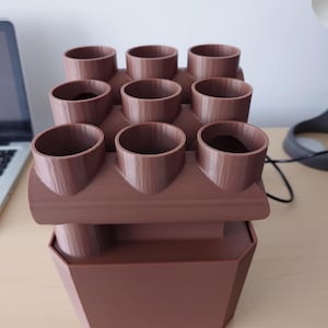 Puede incluir: Un macetero marrón con nueve aberturas cilíndricas dispuestas en tres filas. El macetero tiene una base rectangular. El diseño es moderno y funcional, adecuado para plantas pequeñas o hierbas. El material parece sólido y mate.