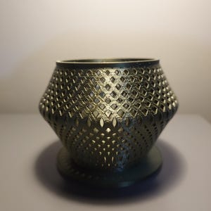 May include: A gold-coloured metal candle holder with a geometric design. The holder has a round base and a wide, open top.