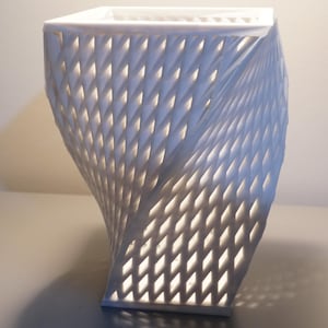 Pode incluir: Objeto decorativo branco e geométrico, com um topo quadrado e um corpo torcido com padrão de diamante. O design permite que a luz filtre, criando sombras. Provavelmente uma lâmpada ou um vaso decorativo.