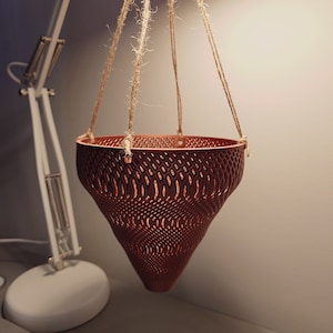 Könnte beinhalten: Ein hängender Pflanzentopf in Rotbraun mit geometrischem Muster, aufgehängt an vier Juteseilen. Der Pflanzentopf hat eine ausgestellte, konische Form. Das Design lässt Licht durchscheinen. Für den Innenbereich.