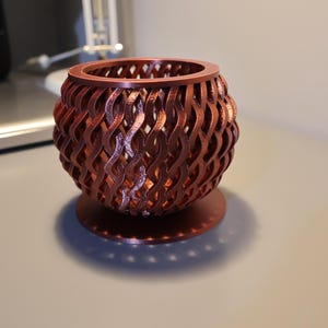 May include: A copper-coloured, 3D printed planter with a geometric, lattice design. The planter has a round base and a round top, and is made of a thin, open-weave material.