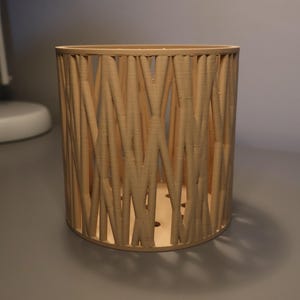 Op de afbeelding: Een cilindrische, lichtbruine decoratieve container met een geweven ontwerp. De container is gemaakt van verticale en diagonale houten latten, waardoor een open, luchtige structuur ontstaat. De basis heeft kleine gaatjes.