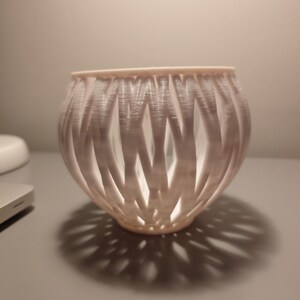 May include: White 3D printed candle holder with a woven design. The holder is round and has a wide opening at the top.