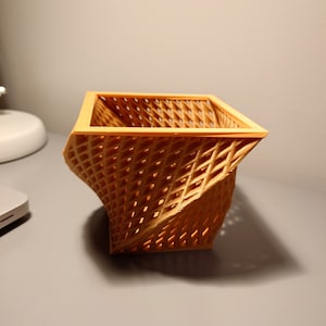 Op de afbeelding: Een beige, 3D-geprinte, geometrische mand met een open geweven structuur, een vierkante basis en een gedraaid, hoekig ontwerp.