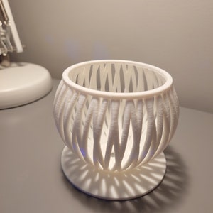 May include: A white, spherical decorative object with a latticework design. The object is made of a series of vertical, curved bars that create an open, airy appearance. It sits on a round base, casting a patterned shadow.