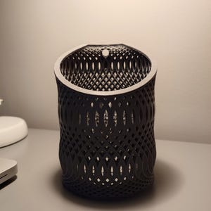 Könnte beinhalten: Ein schwarzer, 3D-gedruckter, zylindrischer Pflanztopf mit geometrischem Muster. Der Topf hat oben ein Loch zum Aufhängen.