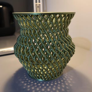 May include: A green, 3D printed vase with a woven, lattice design. The vase is made of plastic and has a cylindrical shape.