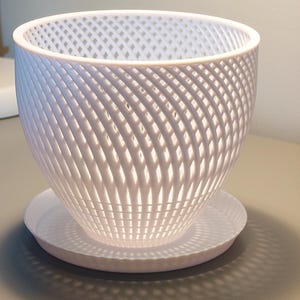 Può includere: Vaso di plastica bianco con un design geometrico a forma di traliccio. Il vaso ha un sottovaso abbinato.