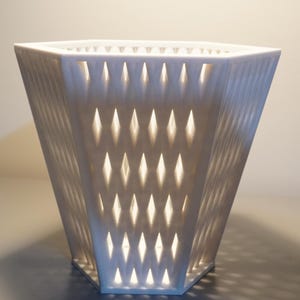 Könnte beinhalten: Ein weißes, geometrisches Dekorationsobjekt mit rautenförmigen Aussparungen. Das Objekt hat eine trapezförmige Form und wird von innen beleuchtet, wodurch Schatten entstehen. Das Design ist modern und minimalistisch, geeignet für die Inneneinrichtung.